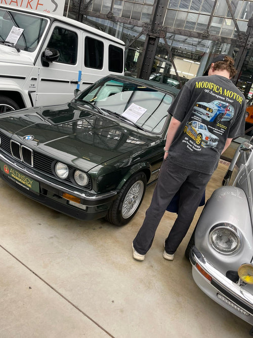 Person wearing a black 90's European Tour T-Shirt | DTM BMW E30 Mercedes 190E Racing Tee with colorful racing car graphics standing next to a classic dark green BMW E30 in a showroom with other vintage cars.