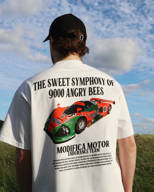 Man wearing a white 787B T-Shirt | Mazda Le Mans 1991 Victory Racing Tee with a colorful race car graphic and bold text on the back standing outdoors under a partly cloudy sky.
